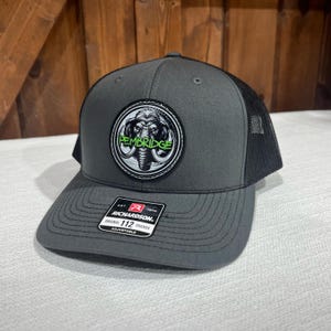 May include: A gray and black trucker hat with a black mesh back. The hat has a round patch with a gray and white elephant graphic and the text "PEMBRIDGE" in green letters.