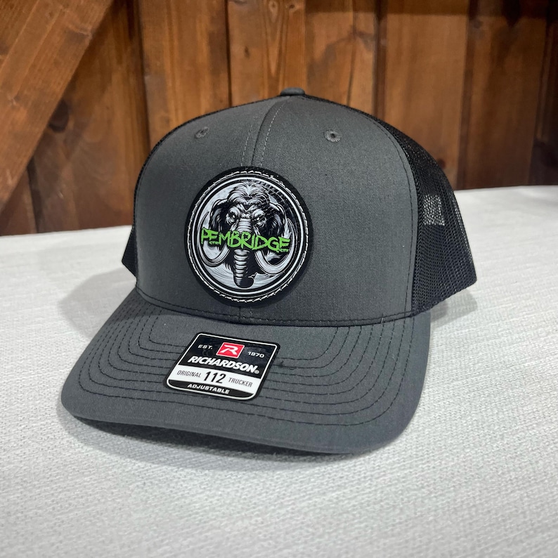 May include: A gray and black trucker hat with a black mesh back. The hat has a round patch with a gray and white elephant graphic and the text "PEMBRIDGE" in green letters.