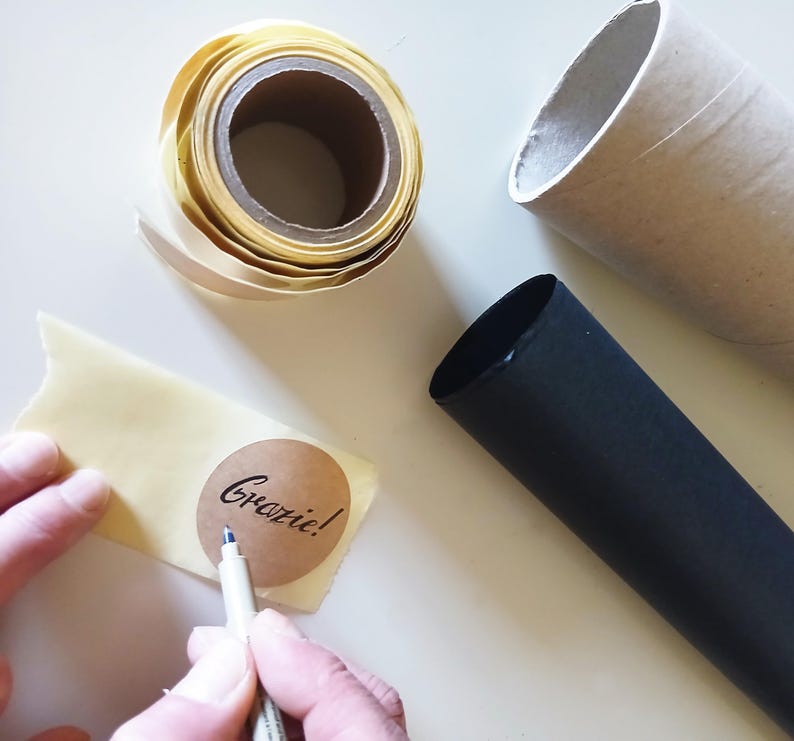 May include: A roll of pale yellow adhesive tape, a brown cardboard tube, a black cylindrical tube, and a circular brown sticker featuring the word Grazie! written in black cursive script, being marked with a fine-tipped grey pen.