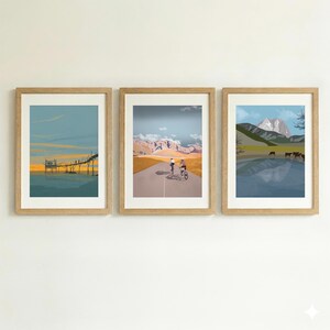 May include: A set of three framed art prints. The left print shows a wooden pier over water at sunset. The center print depicts two cyclists on a road toward mountains. The right print features a lake with cows and a mountain peak in the background.