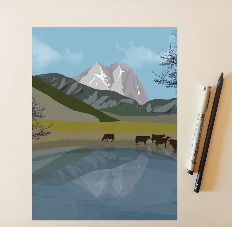 May include: A digital art print depicting a mountain landscape with a lake in the foreground, featuring a group of dark brown cows grazing on the shore. The scene includes a snow-capped peak, rolling hills, and a clear blue sky. Two pens are placed beside the print.