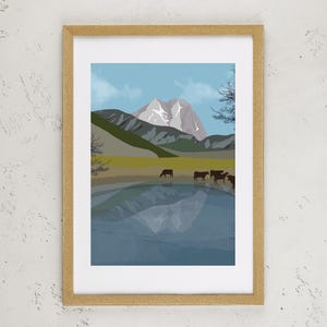 May include: A framed art print featuring a landscape illustration of a snow-capped mountain peak rising above a grassy field with a small herd of dark brown cows standing near the edge of a calm, reflective blue lake under a light blue sky.