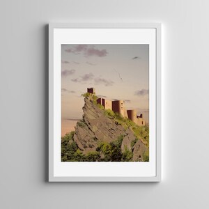 May include: A framed art print featuring a digital illustration of a stone castle perched atop a jagged, rocky cliffside. The scene is set against a soft, pale sky with scattered clouds, with green foliage growing along the base of the mountain.