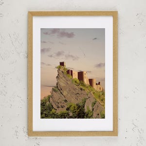 May include: A framed art print featuring a stone castle perched atop a jagged, rocky cliffside. The structure is rendered in warm, earthy tones against a soft, pale sky. The frame is a light-colored wood, surrounding a white mat and the central image.