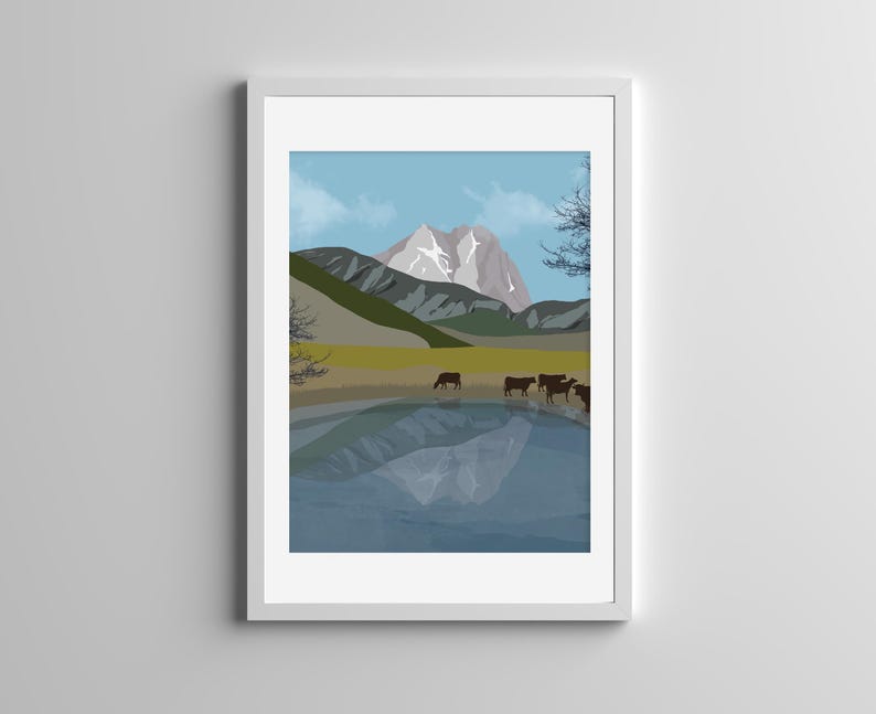 May include: A framed digital art print featuring a mountain landscape with a snow-capped peak, a grassy meadow, and a calm lake reflecting the mountains. A small herd of dark brown cows stands near the water's edge under a light blue sky.