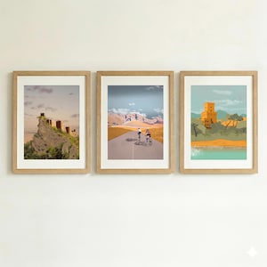 May include: Three framed art prints in light wood frames. The left print shows a stone castle on a rocky cliff. The center print depicts two cyclists on a road toward mountains. The right print features a yellow tower near trees and water.