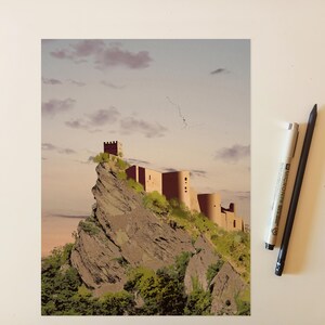 May include: A rectangular art print depicting a castle perched atop a jagged, rocky cliffside with green vegetation, set against a soft sunset sky with scattered clouds. A white fine-liner pen and a black pencil are positioned to the right.