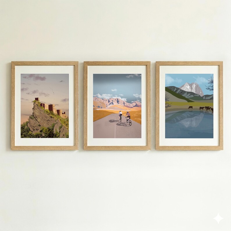 May include: Three framed wall art prints in light wood frames. The left print shows a stone castle on a rocky cliff. The center print depicts two cyclists on a road with mountains. The right print shows a lake with mountains and grazing cattle.