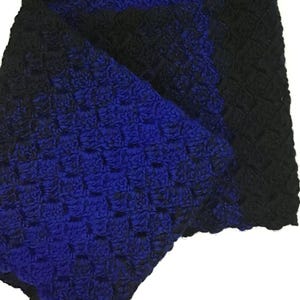 May include: A handmade crochet blanket in shades of blue and black. The blanket features a textured stitch pattern and a scalloped edge. The colors transition from blue to black, creating a gradient effect. The blanket is folded, showing both sides.