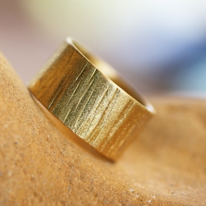 May include: A gold ring with a textured, vertical striped pattern. The ring is lying on a brown surface.