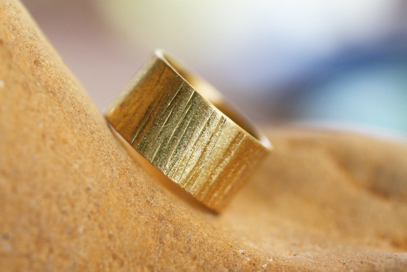 May include: A gold ring with a textured, vertical striped pattern. The ring is lying on a brown surface.