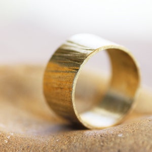 May include: A gold-colored ring with a textured surface. The ring is lying on a brown, textured surface.