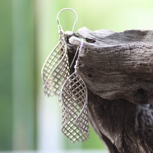 May include: A pair of silver earrings with a geometric, lattice design. The earrings are hanging from a wooden surface.