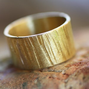 May include: A gold ring with a textured, wood-like finish. The ring is wide and has a rounded shape.