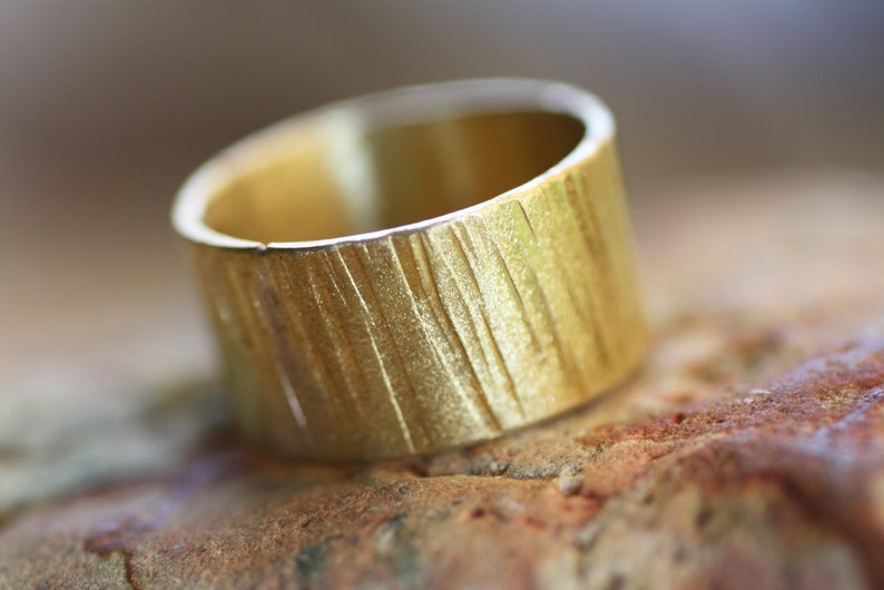 May include: A gold ring with a textured, wood-like finish. The ring is wide and has a rounded shape.