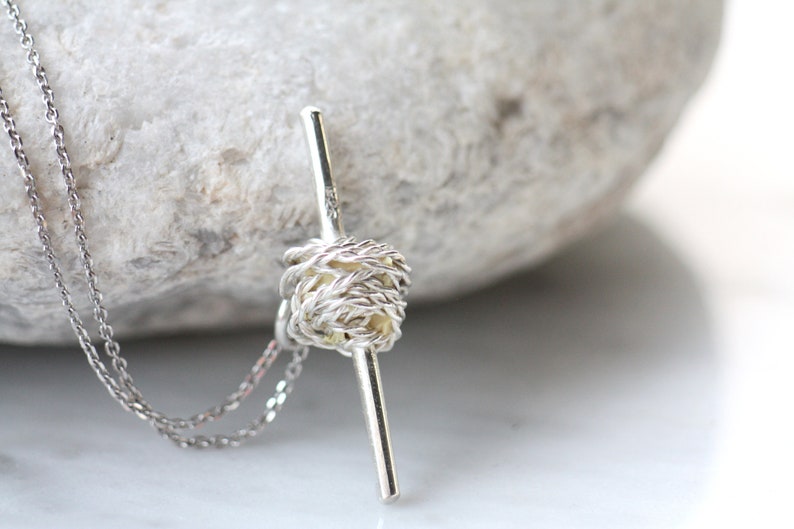 May include: A silver necklace with a knotted pendant and a long, thin bar. The pendant is made of silver wire that is twisted and knotted together to create a unique and intricate design.