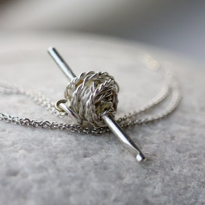 May include: A silver necklace with a yarn ball and knitting needle pendant. The pendant is made of twisted wire and has a realistic look.