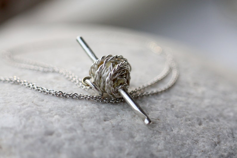 May include: A silver necklace with a yarn ball and knitting needle pendant. The pendant is made of twisted wire and has a realistic look.
