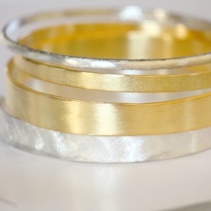 Puede incluir: Cuatro pulseras de oro y plata apiladas una encima de la otra. Las pulseras son de diferentes anchos y tienen un acabado cepillado.