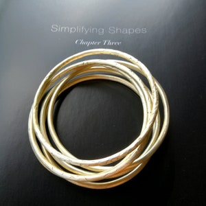 Op de afbeelding: Een set van zes gouden metalen armbanden met een getextureerd oppervlak. De armbanden zijn op elkaar gestapeld.