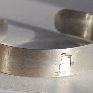 Puede incluir: Un brazalete de plata con un diseño grabado simple de un árbol en la parte delantera.
