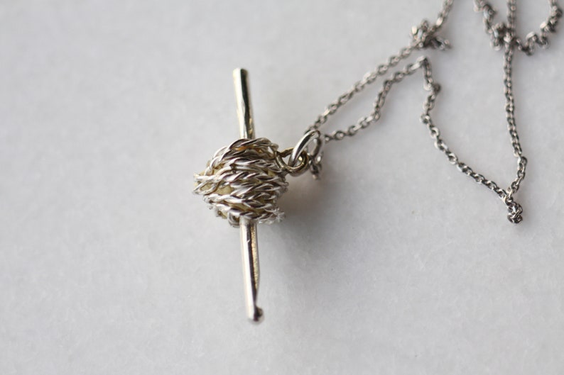 May include: A silver necklace with a yarn ball and crochet hook charm. The charm is detailed and realistic.