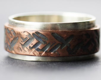 May include: A silver and copper ring with a patterned design. The ring is made of two metals, with the copper band on the outside and the silver band on the inside.