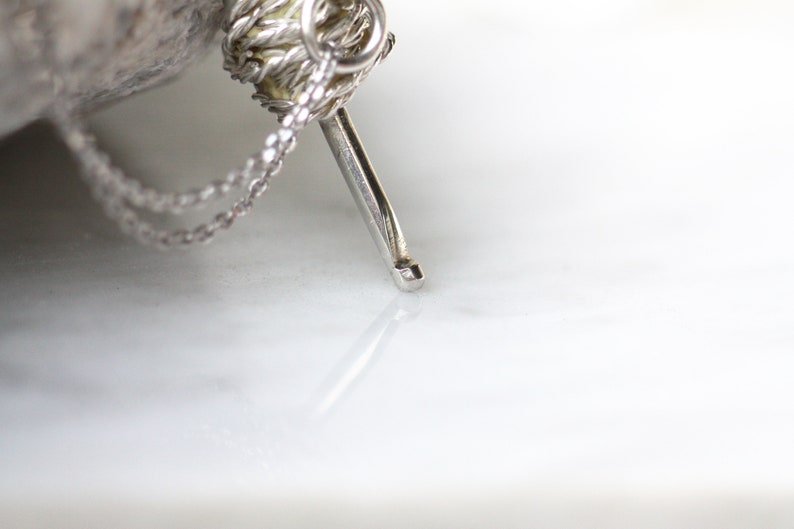 May include: A silver chain necklace with a small, silver, yarn ball charm. The charm has a silver needle attached to it.