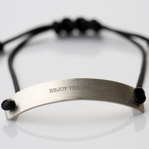 May include: A silver bracelet with a black cord and the words "ENJOY THE JOURNEY" engraved on the metal piece.