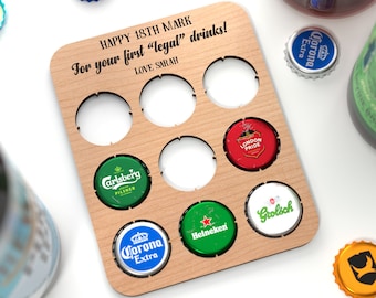 Puede incluir: Tablero de madera con ocho recortes circulares para tapas de botellas. El tablero tiene el texto "HAPPY 18TH MARK For your first "legal" drinks! LOVE SARAH" impreso en él. Hay ocho tapas de botellas en los recortes, incluyendo Corona Extra, Carlsberg, Heineken, Grolsch y London Pride.