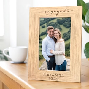 May include: A light-colored wooden picture frame with the word "engaged" in script at the top. The frame holds a photo of a couple and is inscribed with "Mark & Sarah" and the date "12.08.2025". A white mug sits to the left.