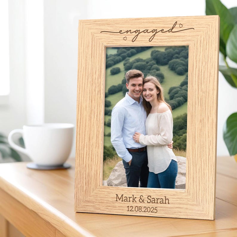 May include: A light-colored wooden picture frame with the word "engaged" in script at the top. The frame holds a photo of a couple and is inscribed with "Mark & Sarah" and the date "12.08.2025". A white mug sits to the left.
