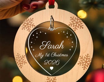 May include: A round wooden Christmas ornament featuring a laser-cut snowflake border and a central clear acrylic heart insert. The heart is engraved with the name Sarah, the text My 1st Christmas 2026, star accents, and a small heart icon.