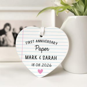 Puede incluir: Adorno de cer&aacute;mica en forma de coraz&oacute;n con dise&ntilde;o de papel rayado. El texto dice "FIRST ANNIVERSARY Paper MARK & SARAH 18.08.2026" con un coraz&oacute;n rosa. Se adjunta una cinta blanca para colgar. Un jarr&oacute;n con flores est&aacute; en el fondo.