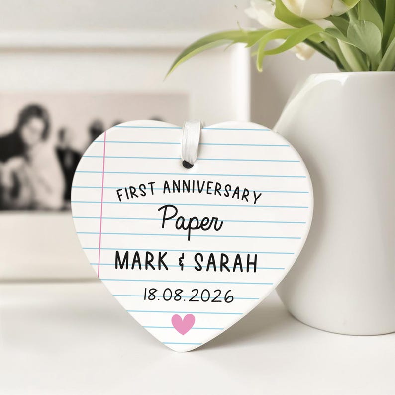 Puede incluir: Adorno de cer&aacute;mica en forma de coraz&oacute;n con dise&ntilde;o de papel rayado. El texto dice "FIRST ANNIVERSARY Paper MARK & SARAH 18.08.2026" con un coraz&oacute;n rosa. Se adjunta una cinta blanca para colgar. Un jarr&oacute;n con flores est&aacute; en el fondo.