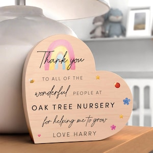 May include: Wooden heart-shaped plaque with a rainbow design and the words "Thank you" and "Oak Tree Nursery." The plaque features decorative bees, stars, and flowers. The message is "for helping me to grow, Love Harry."