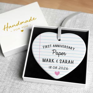 Puede incluir: Adorno en forma de coraz&oacute;n con dise&ntilde;o de papel rayado, con el texto "FIRST ANNIVERSARY Paper MARK & SARAH 18.08.2026" y un coraz&oacute;n rosa. En una caja blanca con la inscripci&oacute;n dorada "Handmade for YOU".