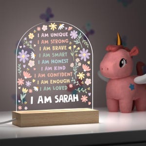 May include: A decorative night light with the text "I AM UNIQUE, I AM STRONG, I AM BRAVE, I AM SMART, I AM HONEST, I AM KIND, I AM CONFIDENT, I AM ENOUGH, I AM LOVED, I AM SARAH" in a floral design. The light sits on a wooden base. A pink unicorn toy is to the right.