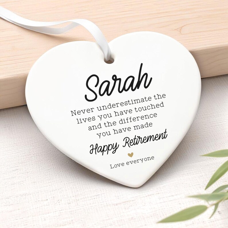 Op de afbeelding: Een wit hartvormig keramisch ornament met de naam "Sarah" in zwart script. Het ornament heeft een wit lint om op te hangen en bevat de tekst "Never underestimate the lives you have touched...Happy Retirement."
