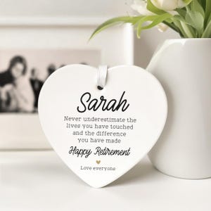 Op de afbeelding: Een witte, hartvormige keramische plaquette met de naam "Sarah" in zwart schrift. De plaquette bevat de tekst "Never underestimate the lives you have touched...Happy Retirement". Een wit lint is bevestigd om op te hangen. Een vaas met bloemen staat op de achtergrond.
