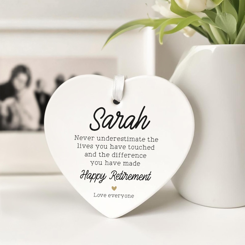 Op de afbeelding: Een witte, hartvormige keramische plaquette met de naam "Sarah" in zwart schrift. De plaquette bevat de tekst "Never underestimate the lives you have touched...Happy Retirement". Een wit lint is bevestigd om op te hangen. Een vaas met bloemen staat op de achtergrond.
