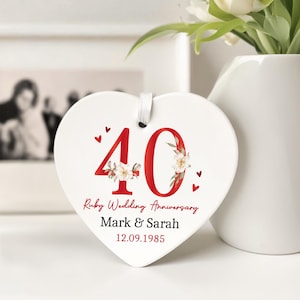May include: A white heart-shaped ceramic ornament with a red '40' and floral details, marking a Ruby Wedding Anniversary. The text reads 'Ruby Wedding Anniversary', 'Mark & Sarah', and the date '12.09.1985'. A white ribbon is attached for hanging.