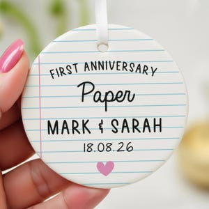 Puede incluir: Adorno redondo de cer&aacute;mica blanca con dise&ntilde;o de papel rayado. El texto dice "FIRST ANNIVERSARY Paper MARK & SARAH 18.08.26" con un coraz&oacute;n rosa en la parte inferior. Una cinta blanca est&aacute; adjunta para colgar.