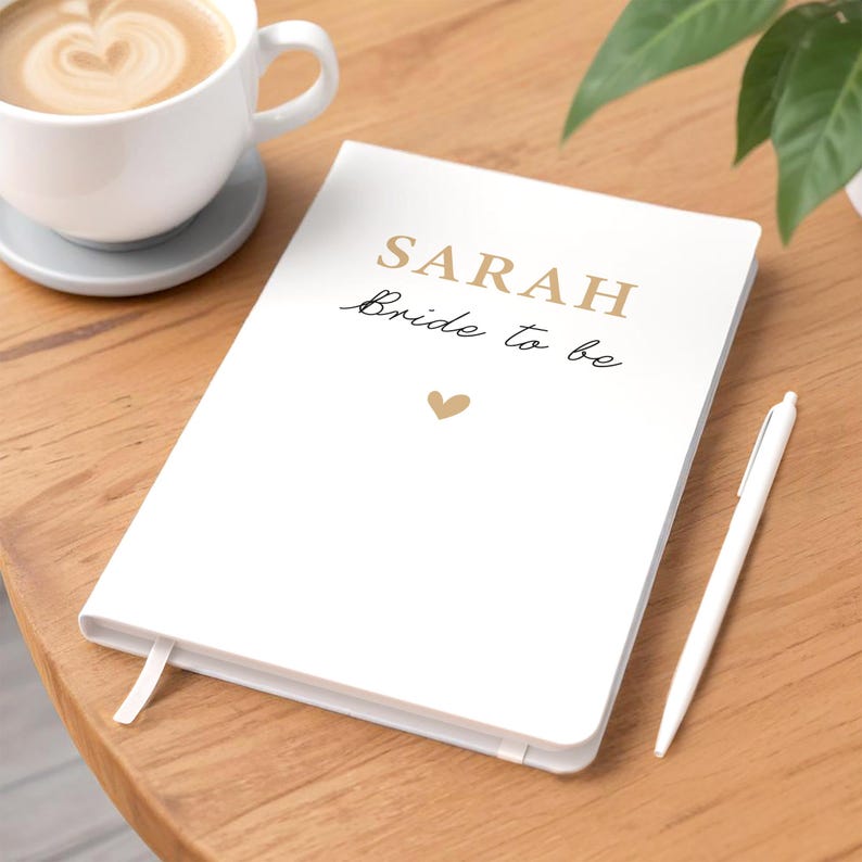 Może przedstawiać: Biały notatnik z imieniem "SARAH" i frazą "Bride to be" wydrukowanymi złotą i czarną czcionką. Złote serce znajduje się pod tekstem. Biały długopis spoczywa obok notatnika na drewnianej powierzchni. Filiżanka kawy jest w tle.