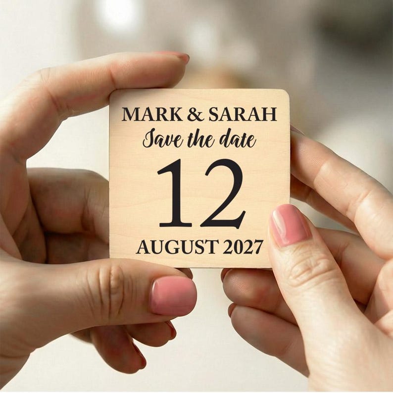 May include: A square wooden save-the-date announcement held by hands. The text reads "MARK & SARAH Save the date 12 AUGUST 2027" in black lettering on a light wood grain background. The hands have pink nail polish.