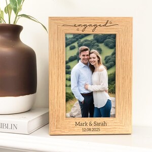 May include: A light-colored wooden picture frame with the word "engaged" in cursive script at the top. The frame holds a photo of a couple and is inscribed with the names "Mark & Sarah" and the date "12.08.2025".