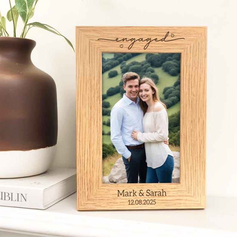 May include: A light-colored wooden picture frame with the word "engaged" in cursive script at the top. The frame holds a photo of a couple and is inscribed with the names "Mark & Sarah" and the date "12.08.2025".