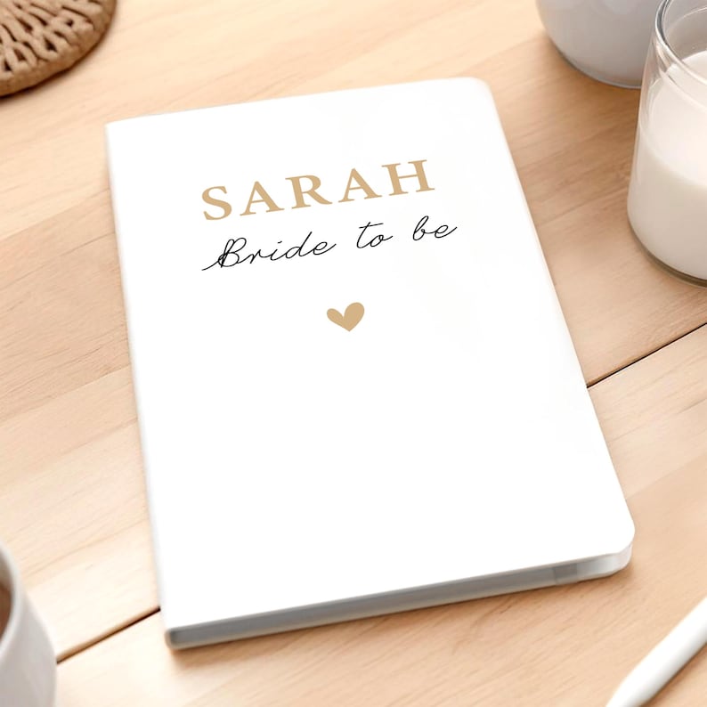 Może przedstawiać: Biały notatnik z napisem "SARAH" złotymi wielkimi literami i "Bride to be" kursywą. Małe złote serce znajduje się pod tekstem. Notatnik leży na jasnej drewnianej powierzchni.