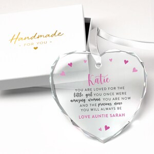 Puede incluir: Un adorno de cristal transparente en forma de coraz&oacute;n con una cinta blanca, presentado en una caja de regalo blanca. El adorno tiene corazones rosas y el texto "Katie, You are loved...Love Auntie Sarah". La caja dice "Handmade for you".