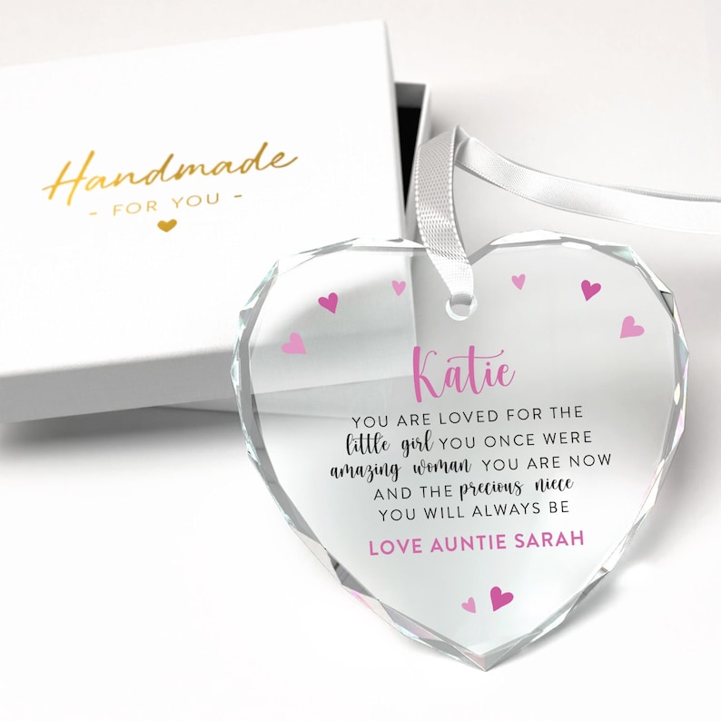 Puede incluir: Un adorno de cristal transparente en forma de coraz&oacute;n con una cinta blanca, presentado en una caja de regalo blanca. El adorno tiene corazones rosas y el texto "Katie, You are loved...Love Auntie Sarah". La caja dice "Handmade for you".
